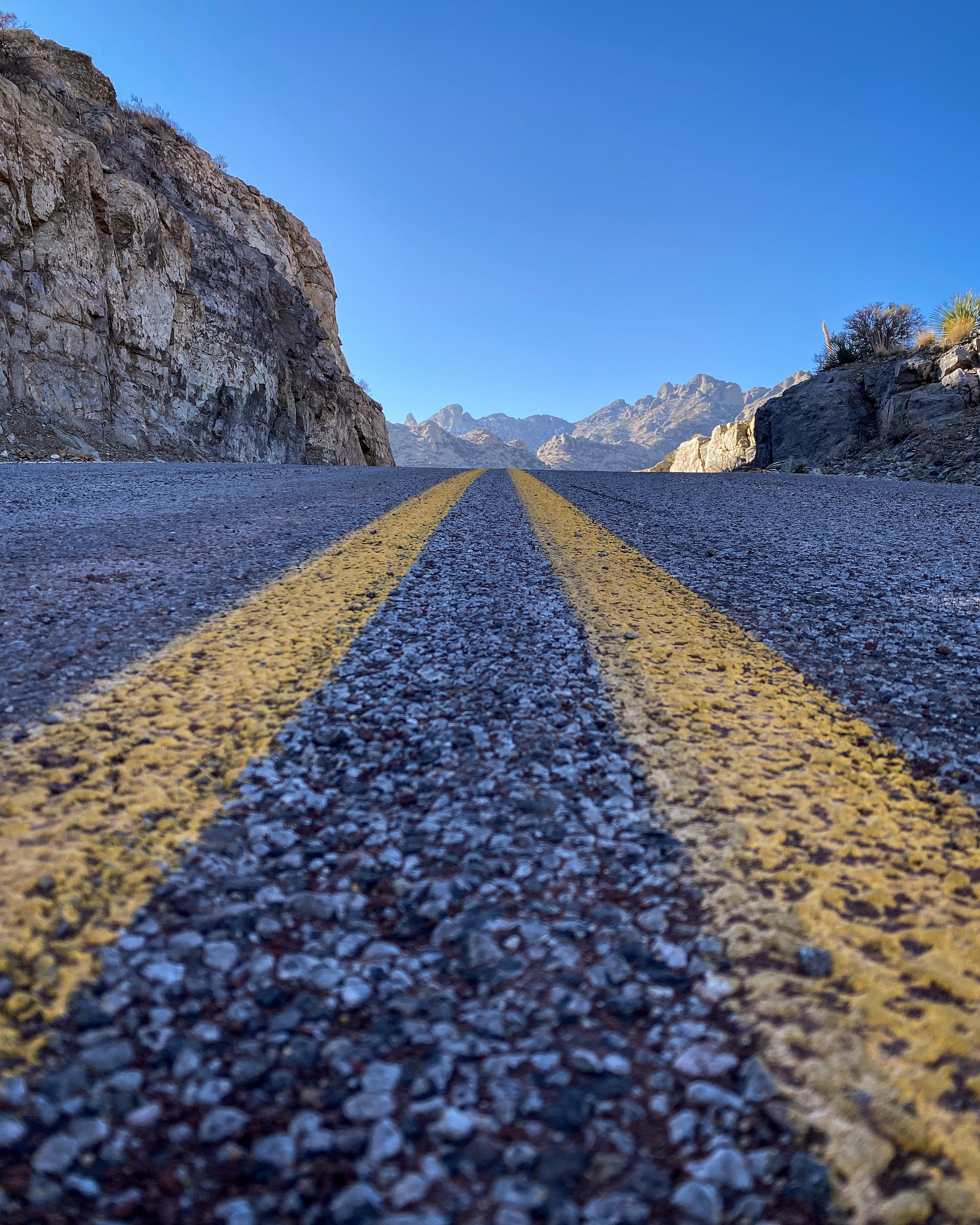 Mountain Road Double yellow road lines leading between rock walls toward mountains beneath a blue sky.